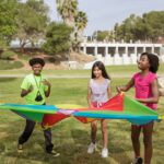 25 juegos cooperativos y de expresión corporal para Infantil y Primaria | Guía práctica Juegos cooperativos: grupo de niños jugando con un paracaídas de colores